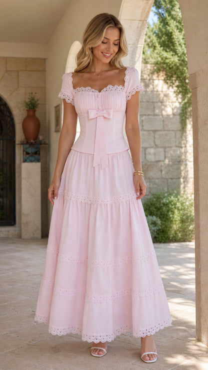 Front full-length view of model wearing a blush pink lace-trimmed tiered maxi dress with bow waist detail