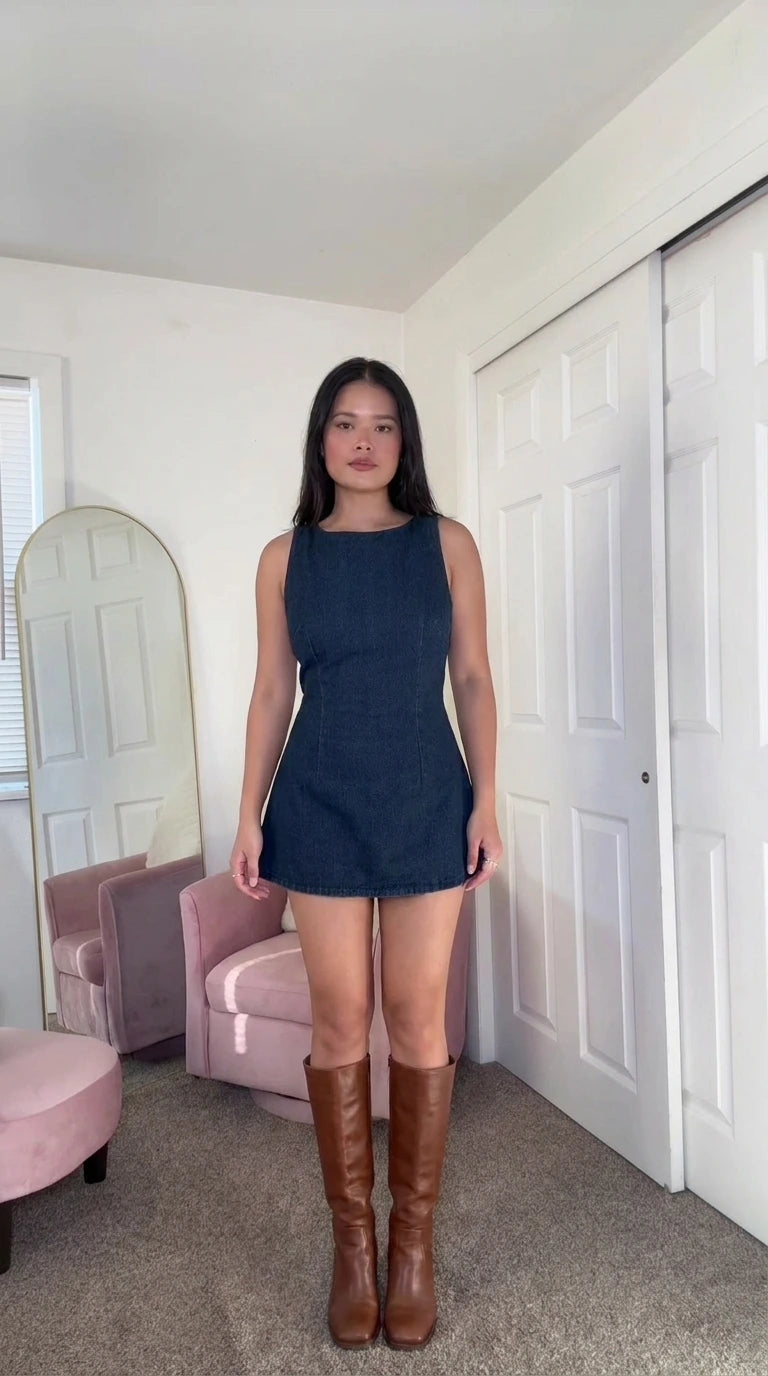 Front view of woman wearing dark blue sleeveless denim mini dress with tall brown boots