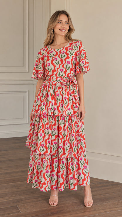 Front full-length view of woman wearing red floral wave print tiered maxi dress with tie waist