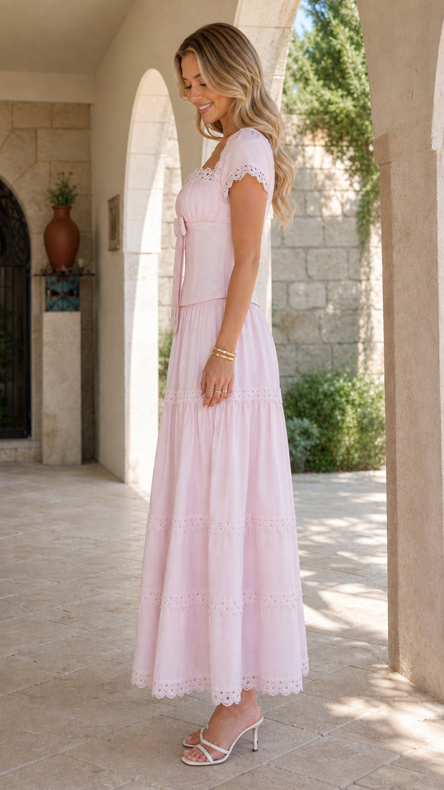Side full-length view of model wearing a blush pink romantic tiered maxi dress in an outdoor courtyard