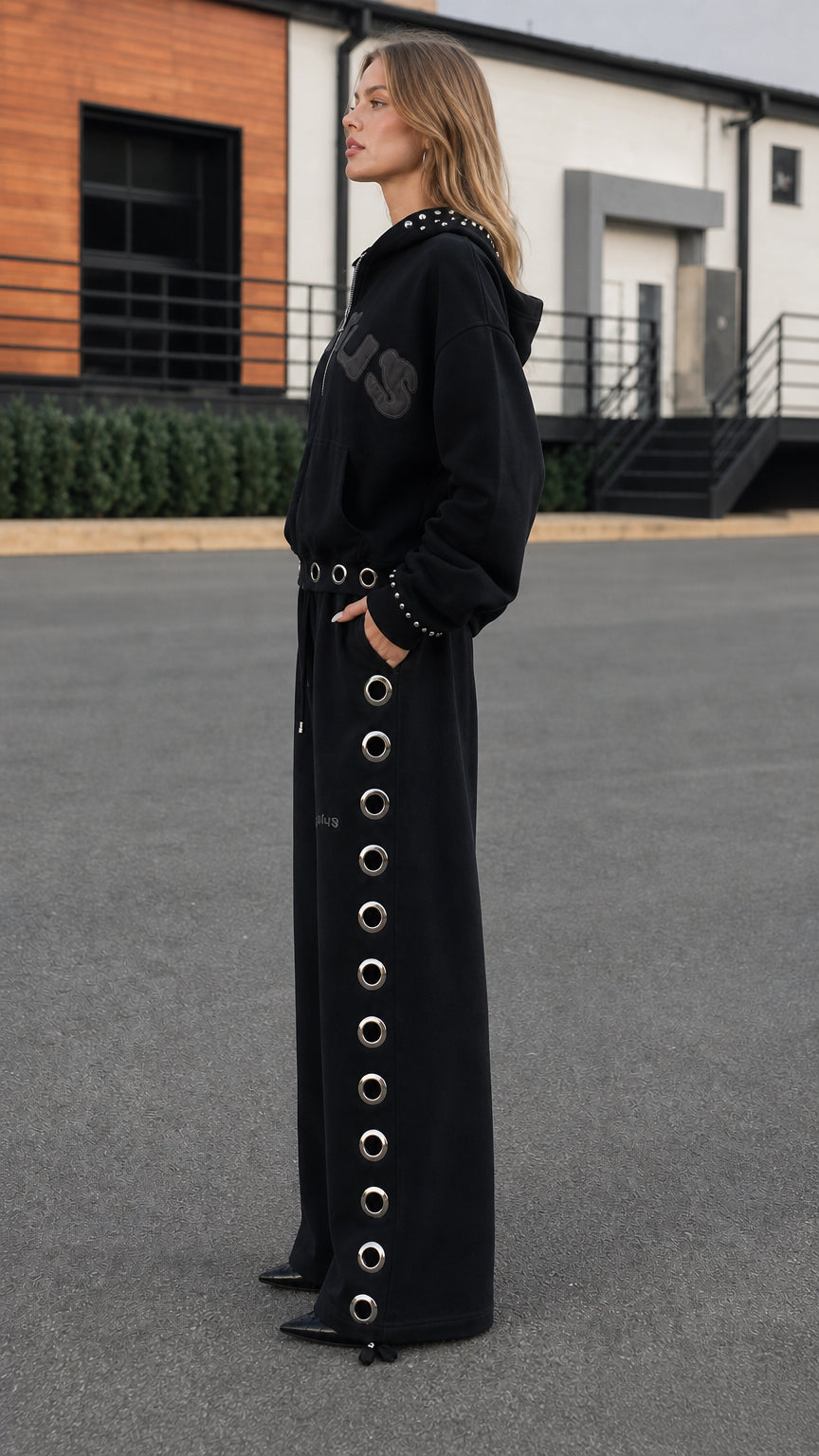 Side profile view of model wearing black studded hoodie and wide-leg eyelet sweatpants