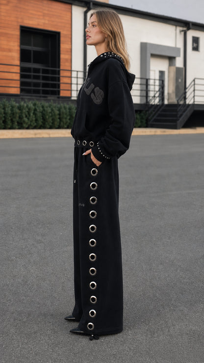 Side profile view of model wearing black studded hoodie and wide-leg eyelet sweatpants