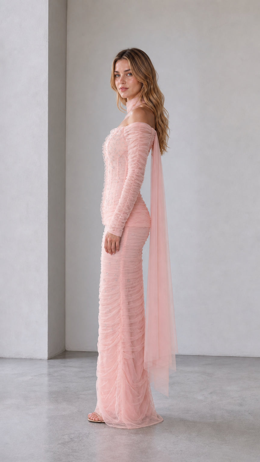 Blush pink fitted evening gown shown from the side with flowing tulle overlay