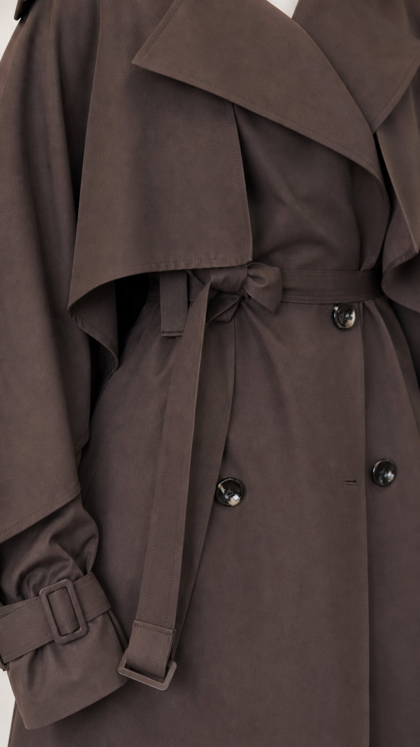 Close-up of dark brown trench coat cape overlay, belt buckle, cuff strap, and buttons