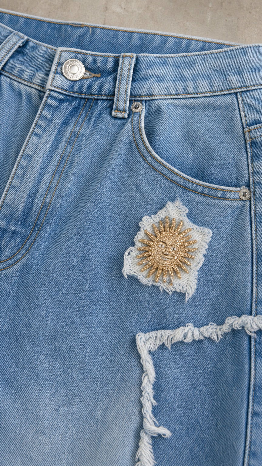 Close-up detail of light blue denim showing raw distressed star patch and golden sun embroidery